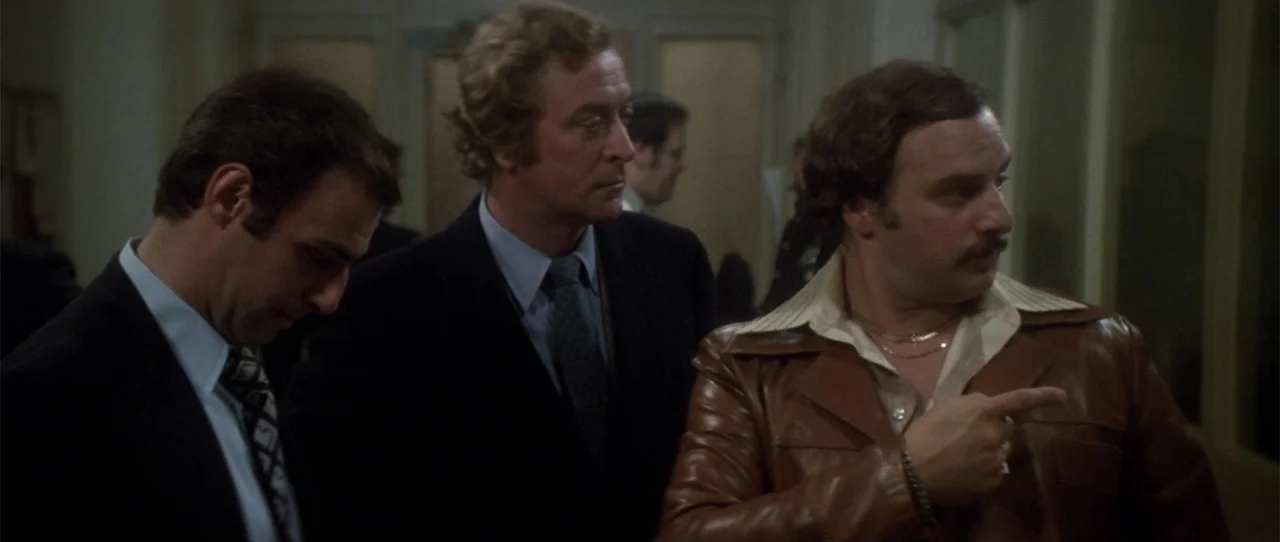 Dressed to Kill (1980) movie still: medium — A 1970s indoor corridor with light-colored walls and doors in the background. Three men stand together…