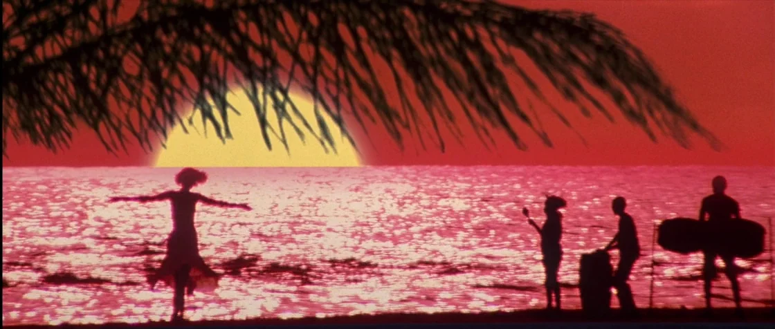 Carlito’s Way (1993) movie still: wide — A beach shoreline at sunset features a palm frond silhouette in the foreground. Four figures appear as dark…