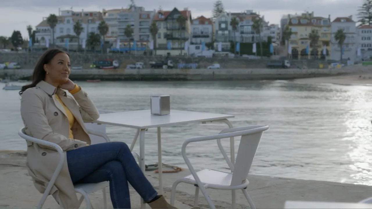 A Pinch of Portugal (2023) movie still: medium — A waterfront promenade with a small outdoor cafe table and an urban coastal background. A woman in…
