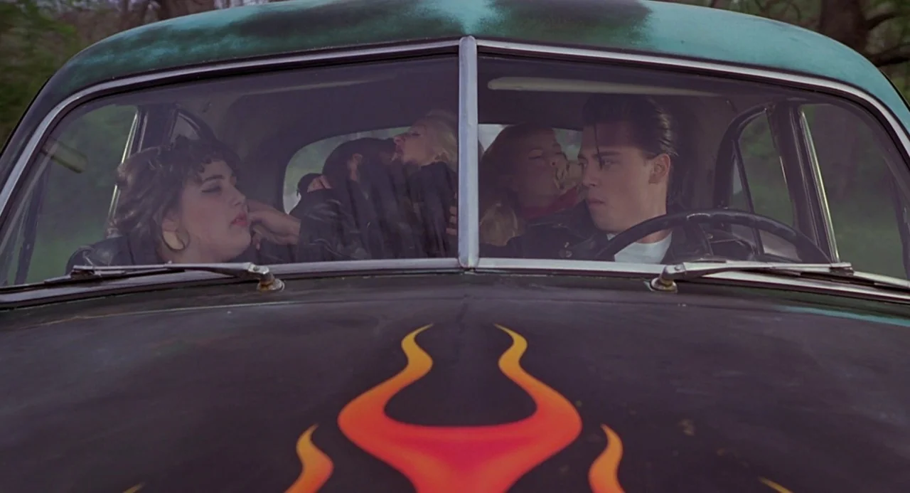 Cry Baby (1990) movie still: medium — Four teenagers are seated inside a vintage car with a distinctive flame-painted hood. The vehicle is parked…