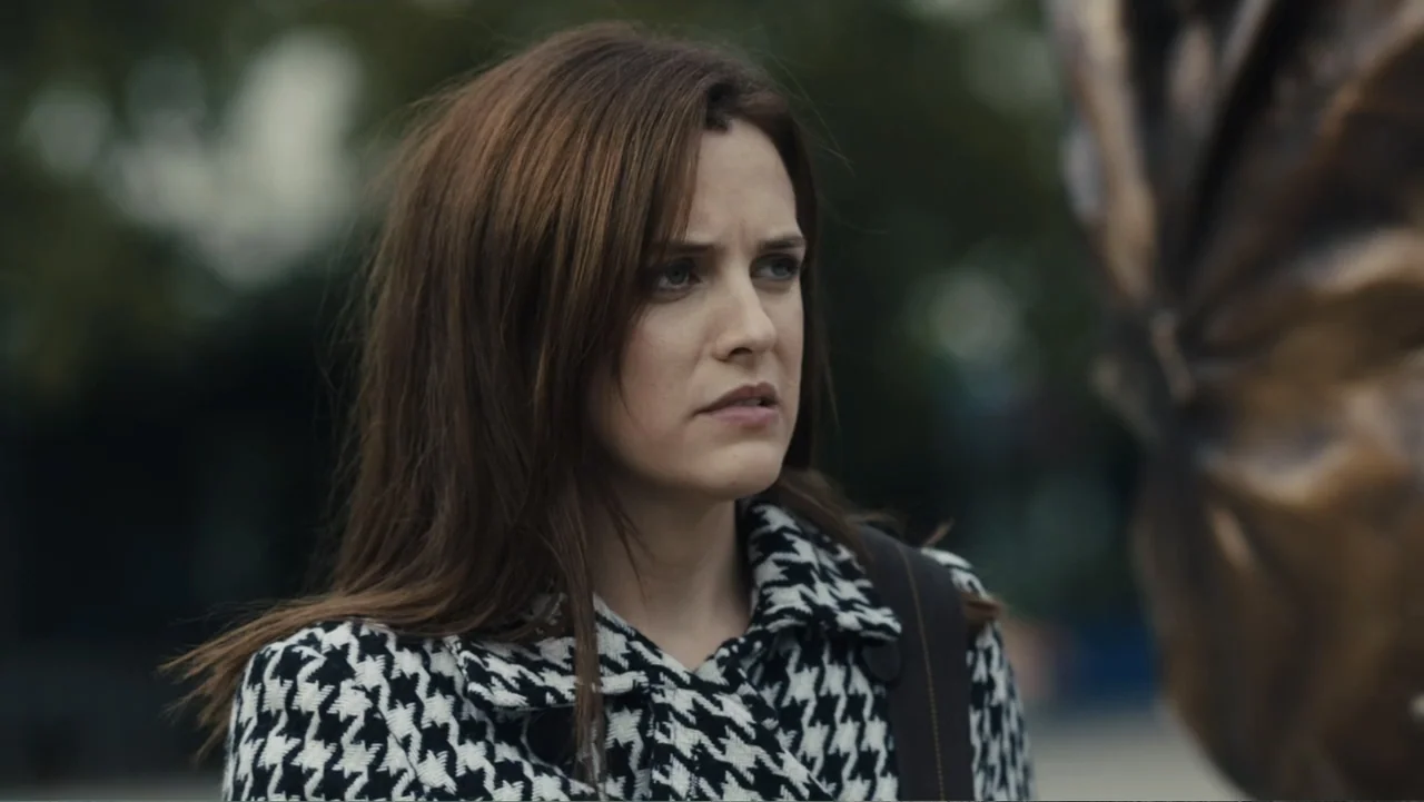 Paterno (2018) movie still: closeup — An outdoor park setting with a blurred background of trees and foliage. A woman in a patterned coat gazes at a…