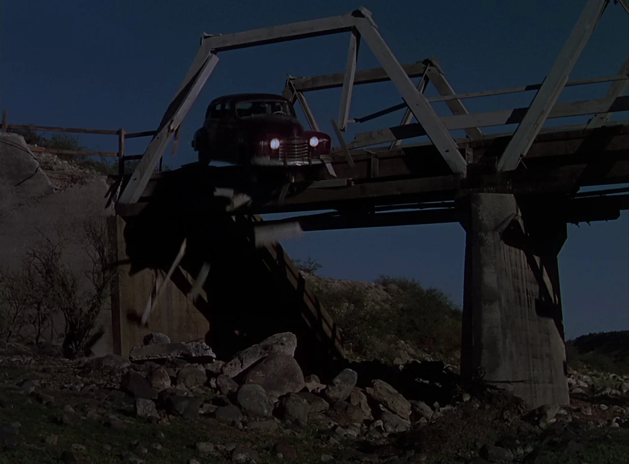 Desert Fury (1947) movie still: wide — A rural wooden bridge partially collapses over a rocky, dry creek bed under daylight. A dark vintage sedan…