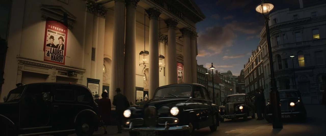 Domino (2005) movie still: wide — A classical theater facade stands on a city street during the twilight hours. Several figures walk toward the…