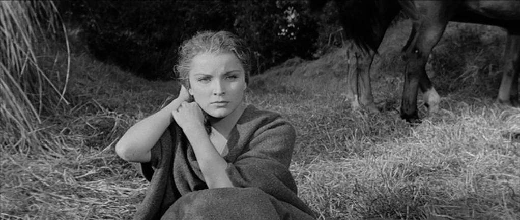 Love Me Tender movie still: medium — A woman sits in a dry, grassy field during the day. A horse stands partially visible in the background next to…