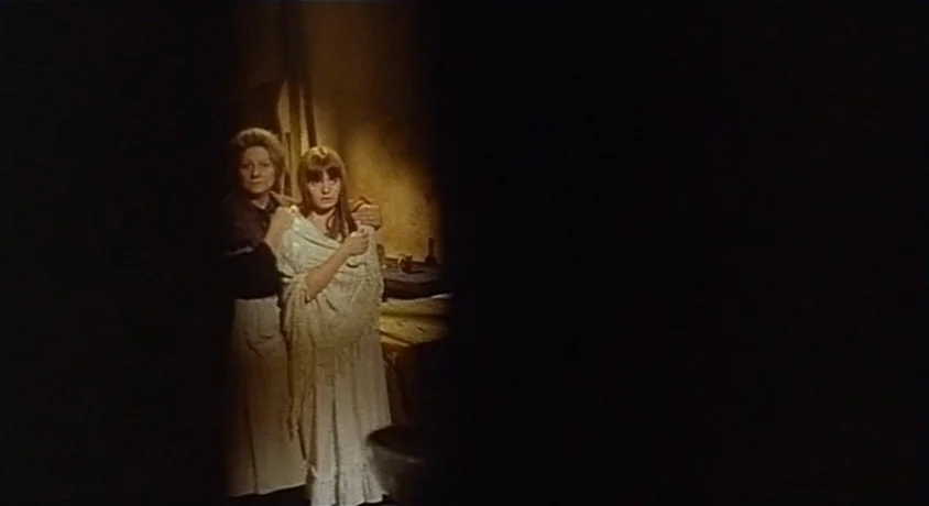 Kill, Baby Kill (1966) movie still: medium — A dimly lit room with textured walls holds two women in period clothing. The younger woman stands with…