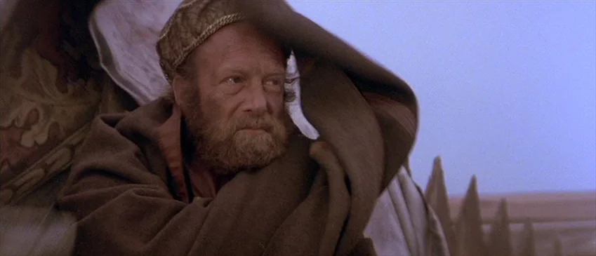 Flesh+Blood (1985) movie still: medium — A man stands in an arid desert landscape with a muted blue sky background. The man has a thick beard and…