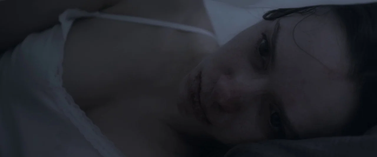 Starry Eyes (2014) movie still: closeup — A person lies under white bedding in a dark, shadow-filled environment. The face is partially lit while…