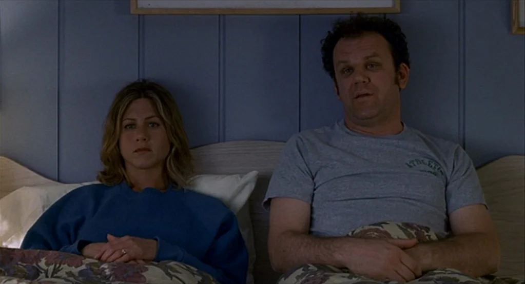 The Good Girl (2002) movie still: medium — A bedroom features blue wood-paneled walls and a headboard behind two people sitting upright. A man and a…
