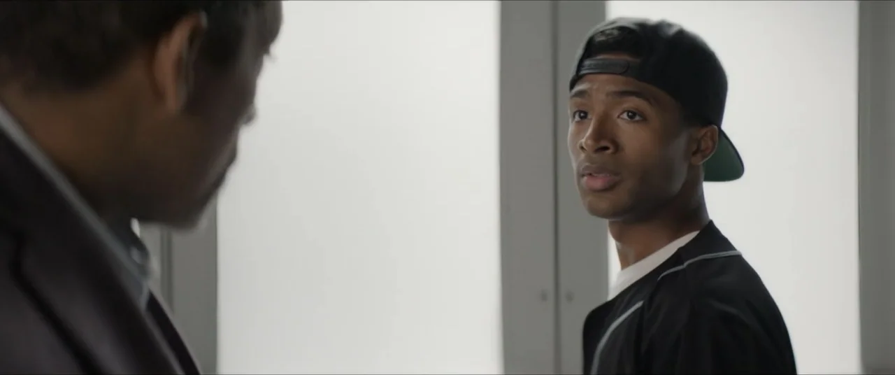 The Forge (2024) movie still: medium — A bright interior space features large white windows providing diffuse natural backlight. A young man wearing…