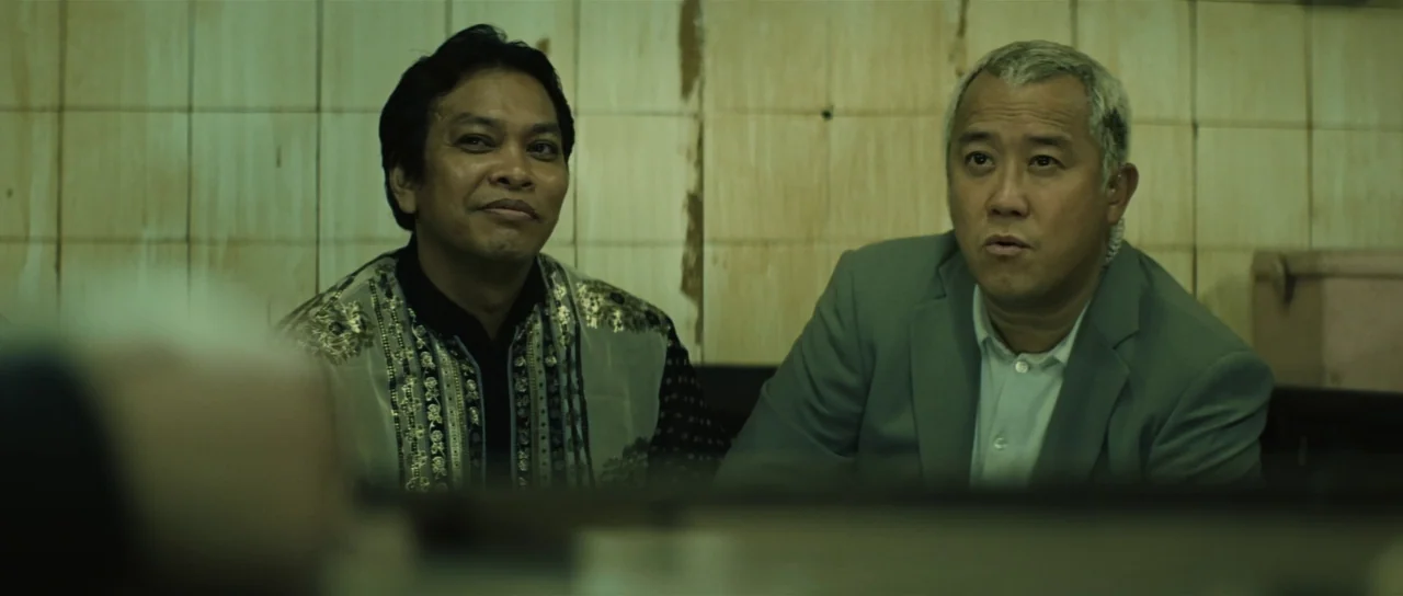 Infernal Affairs (2002) movie still: medium — Two men sit side by side against a tiled wall with visible dirt stains. The men look toward the…