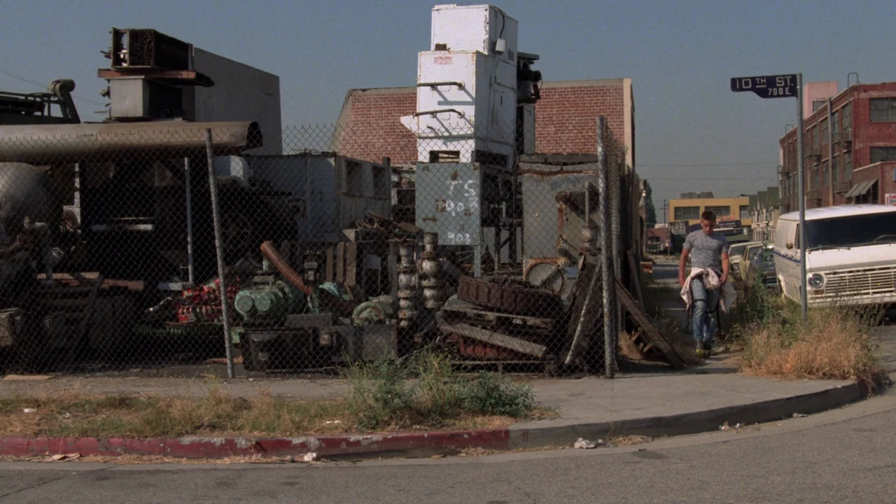 Repo Man (1984) movie still: wide — A fenced scrap metal lot sits on a street corner under bright daylight. A young man walks along the sidewalk…