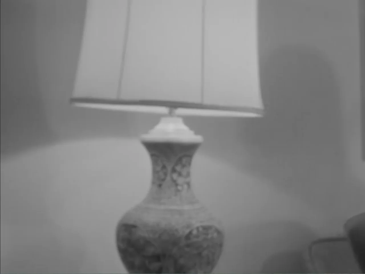 Chained Girls (1965) movie still: closeup — A decorative table lamp with a patterned base stands on a flat surface. The lampshade glows from a light…