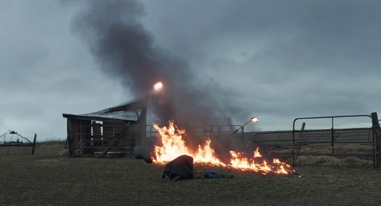 Eternals (2021) movie still: wide — A rural farmyard features a small wooden shed and a metal gate under overcast skies. A person kneels beside…