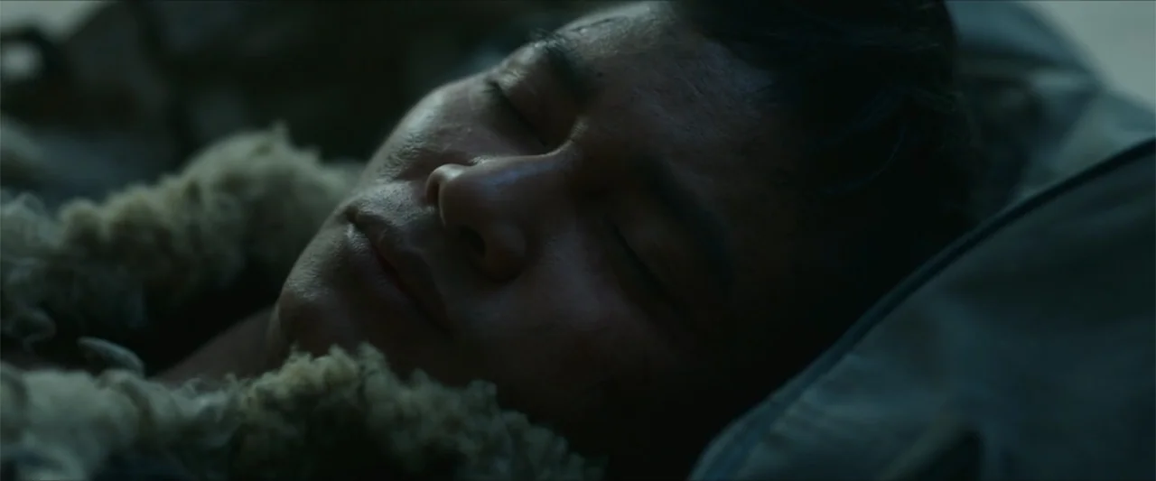 Monster Hunter (2020) movie still: closeup — A dark indoor space with minimal visible detail surrounding the subject. A young man with eyes closed…