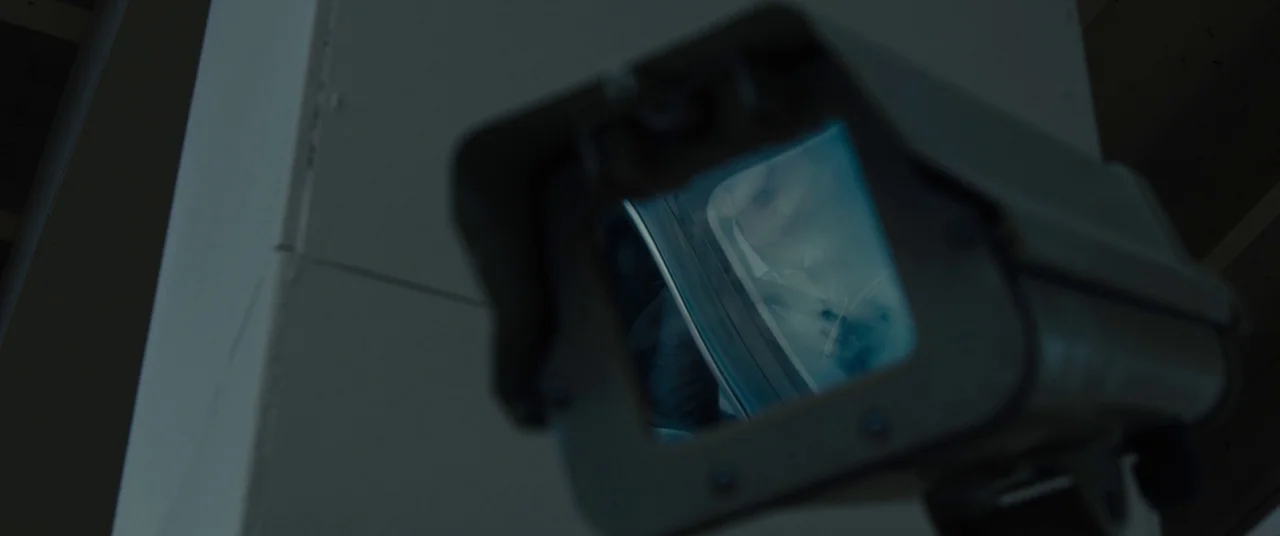 Sicario (2015) movie still: closeup — A close-up shot focuses on a dark gray outdoor security camera housing. The glass lens reflects a blurry,…
