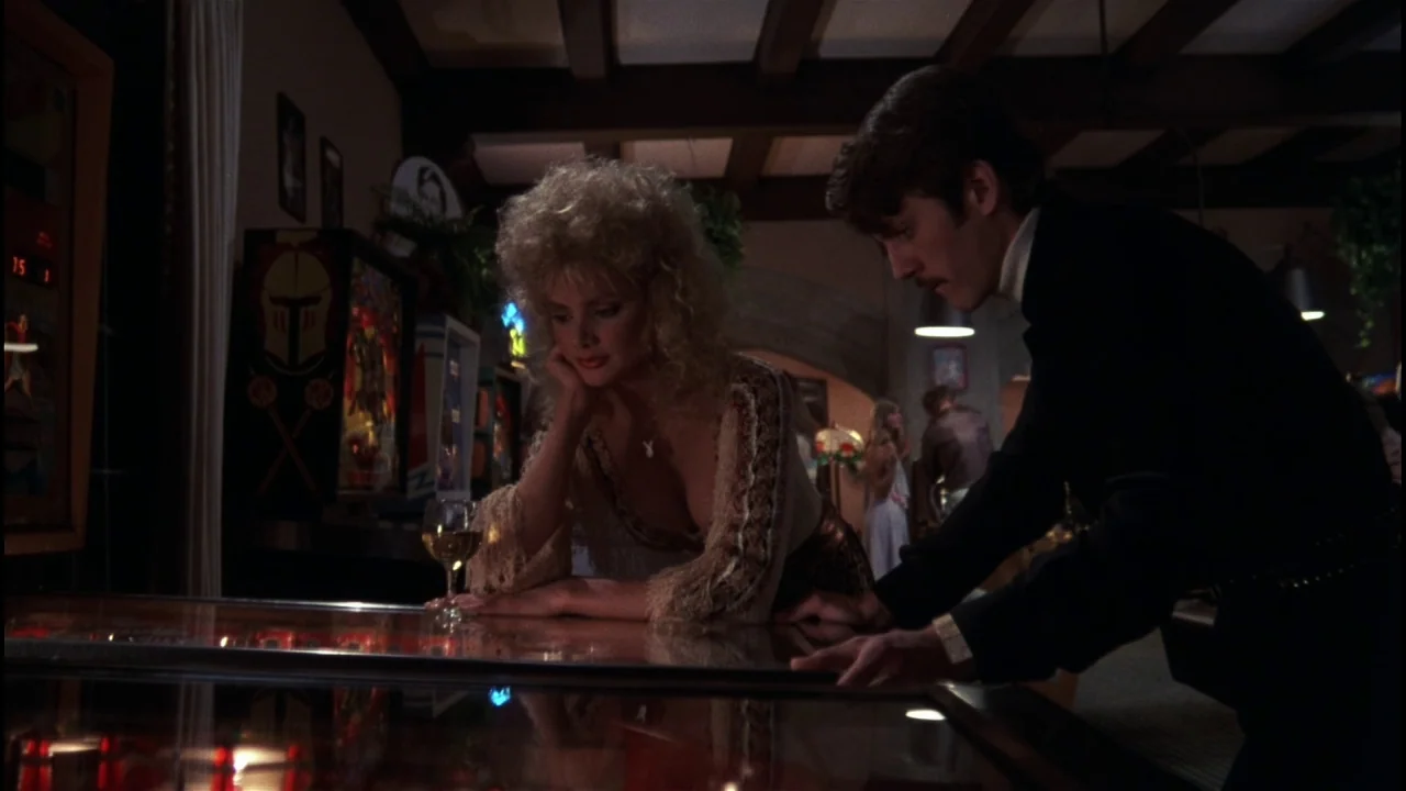 Star 80 (1983) movie still: medium — A dimly lit arcade interior features wooden ceiling beams and rows of upright cabinets. A man and a blonde…