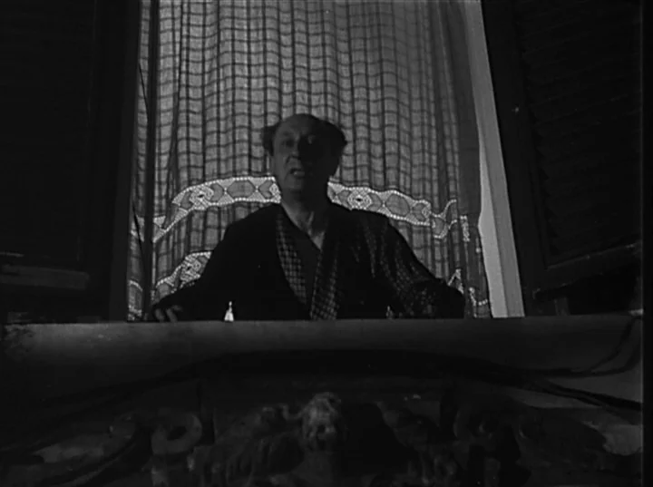 Variety Lights (1950) movie still: medium — A man stands before a curtained window at night. He is positioned in front of a dark, decorative object…