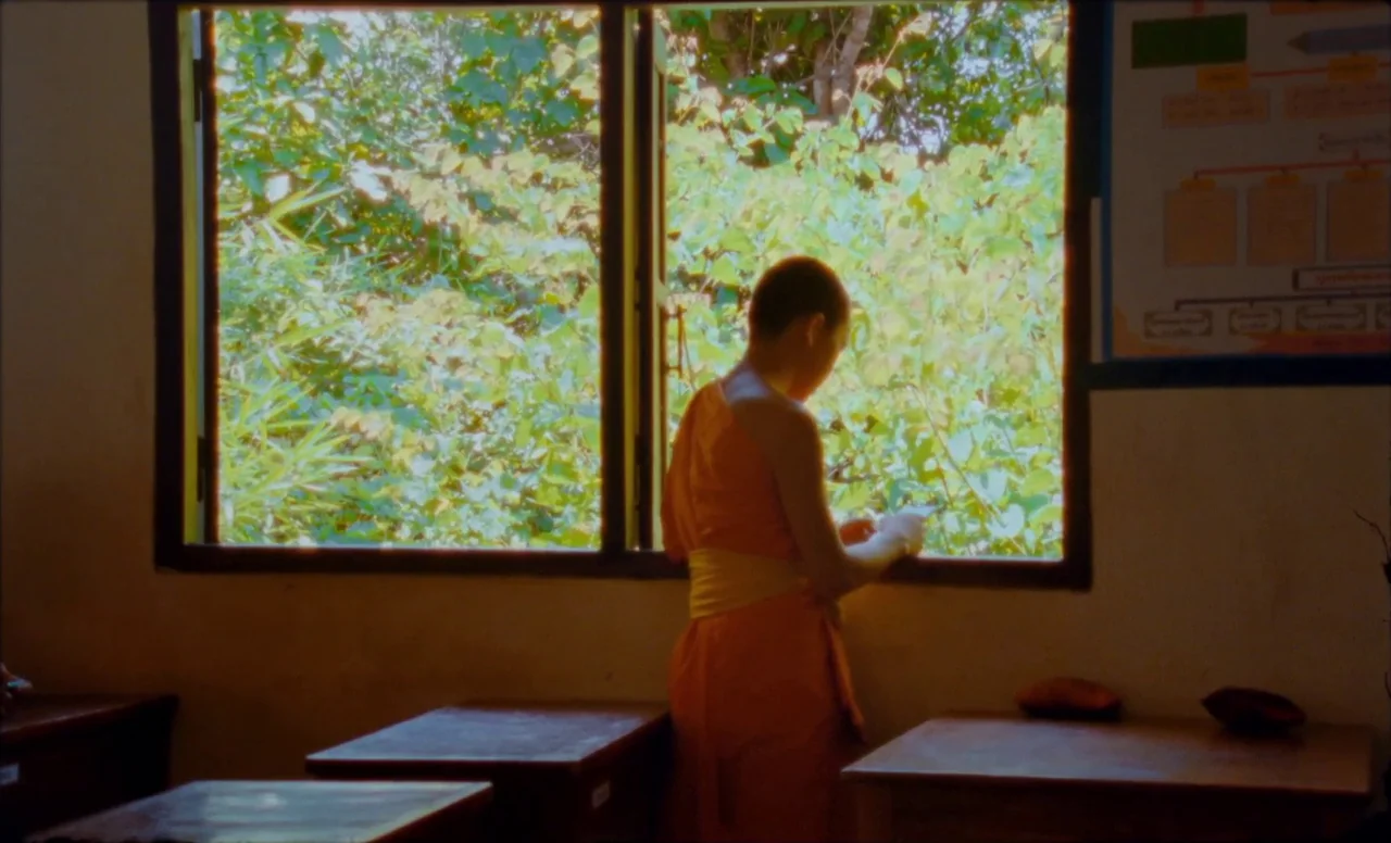 Samsara (2023) movie still: medium — A classroom interior features wooden desks and a large open window overlooking leafy trees. A young monk in…