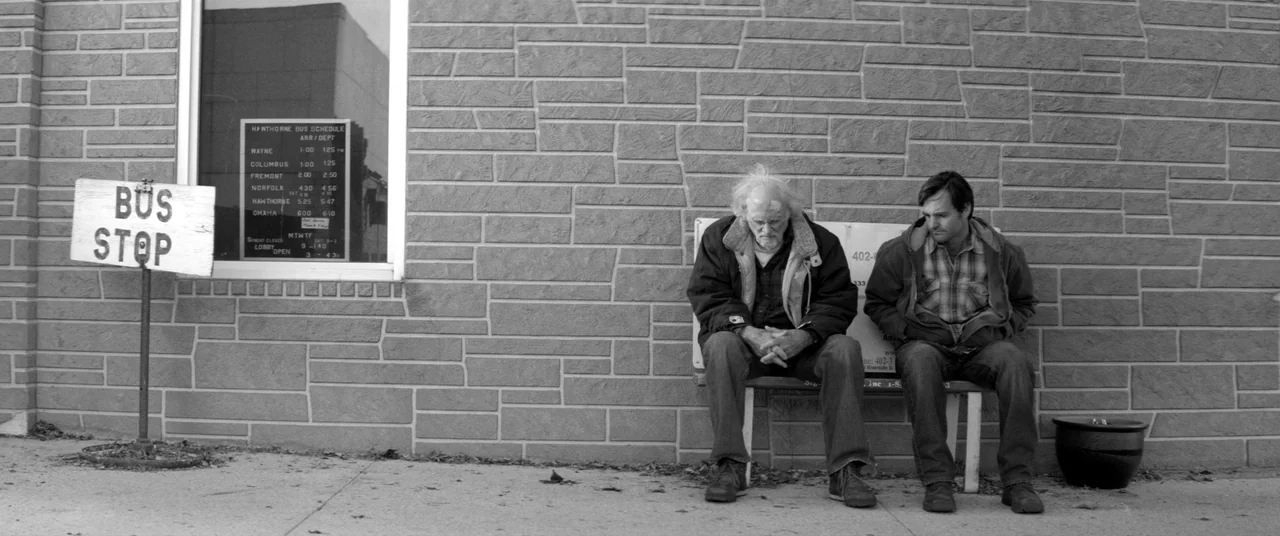 Nebraska (2013) movie still: wide — A bus stop sits against a textured stone wall with a posted schedule. Two men sit side by side on a metal bench…