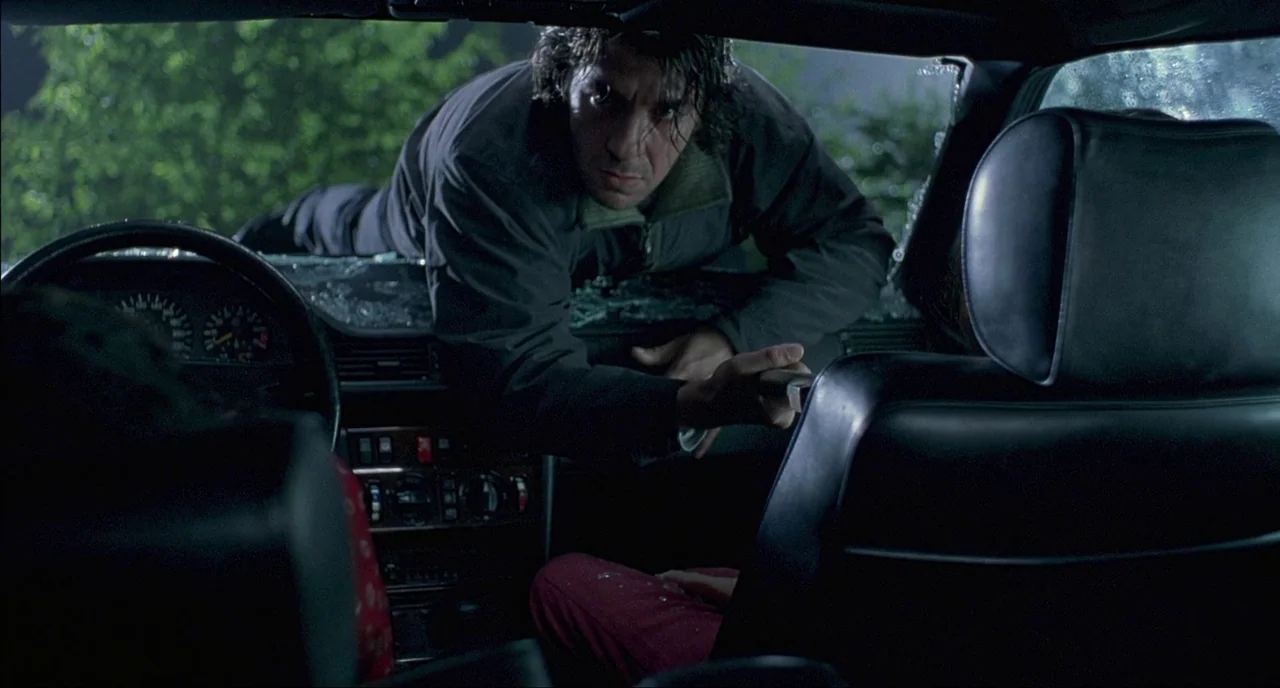 Fat Girl (2001) movie still: medium — A man leans through the shattered windshield of a vehicle parked at night. He reaches into the cabin while the…