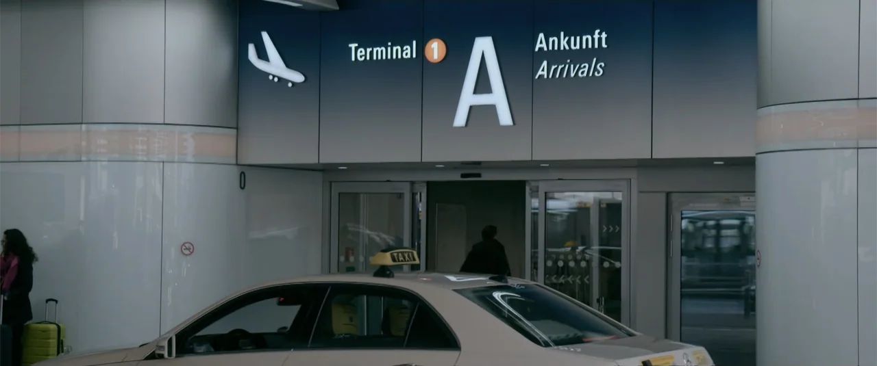 Simple as Water (2021) movie still: medium — An airport arrival area features modern signage above automatic glass sliding doors. A man walks toward…