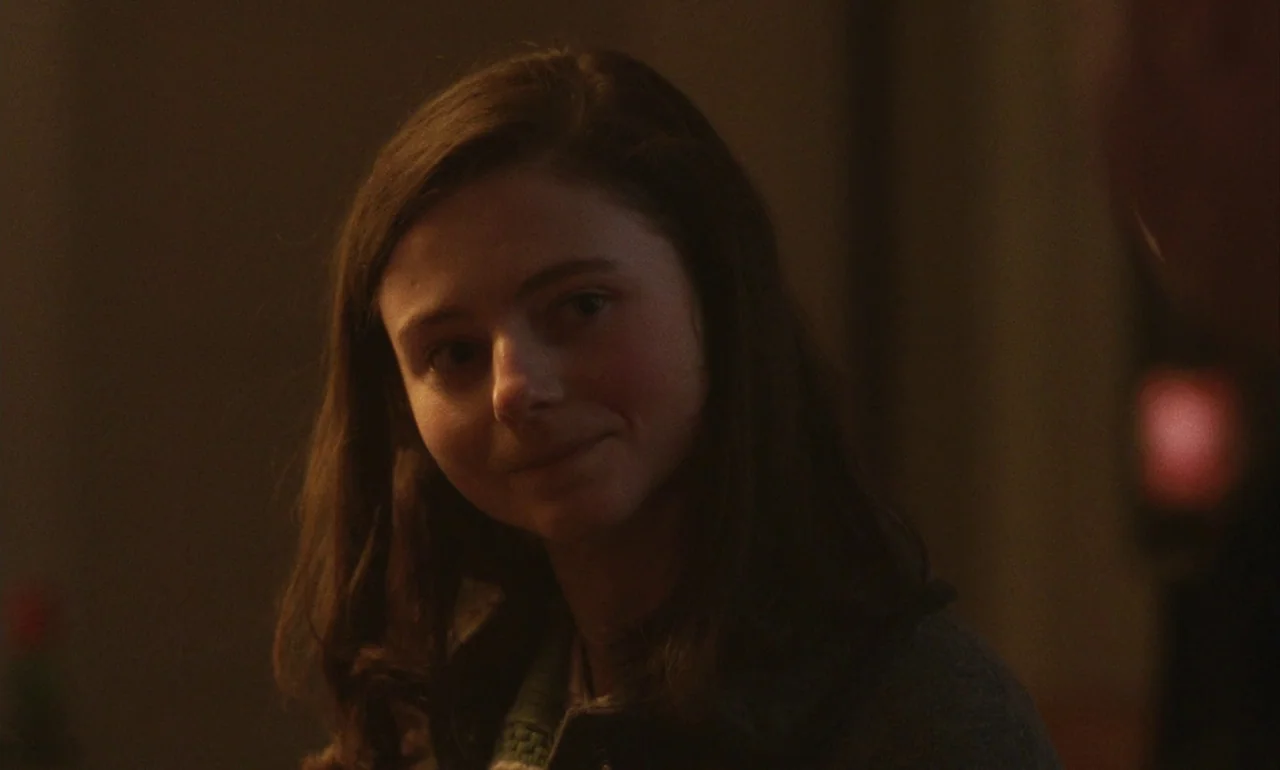 Eileen (2023) movie still: closeup — A dimly lit indoor space with low-contrast, warm shadow tones. A teenage girl looks off-camera with a gentle…