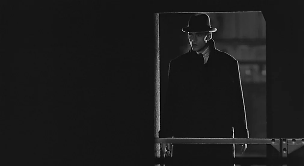 Kafka (1991) movie still: fullbody — A man wearing a hat and coat stands within a dark doorway. He faces the camera while the surrounding…