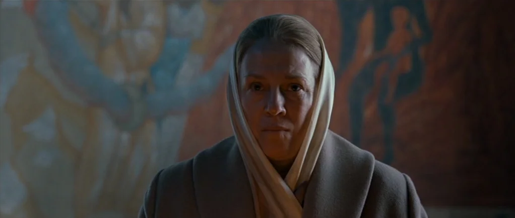 Elena (2011) movie still: closeup — A woman stands before a wall covered by an abstract orange and brown mural. She stares ahead while wearing a…