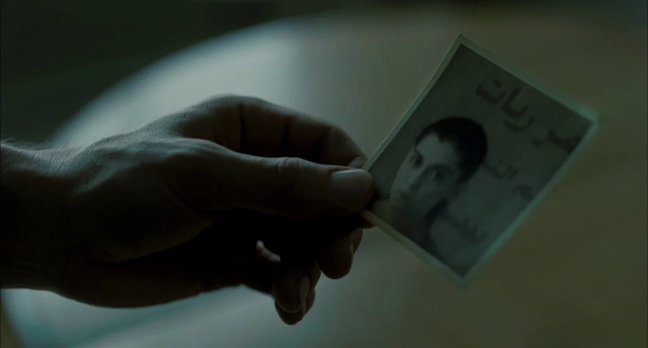 Incendies movie still: closeup — A hand holds a small black and white square photograph with printed Arabic text. The background is a soft,…