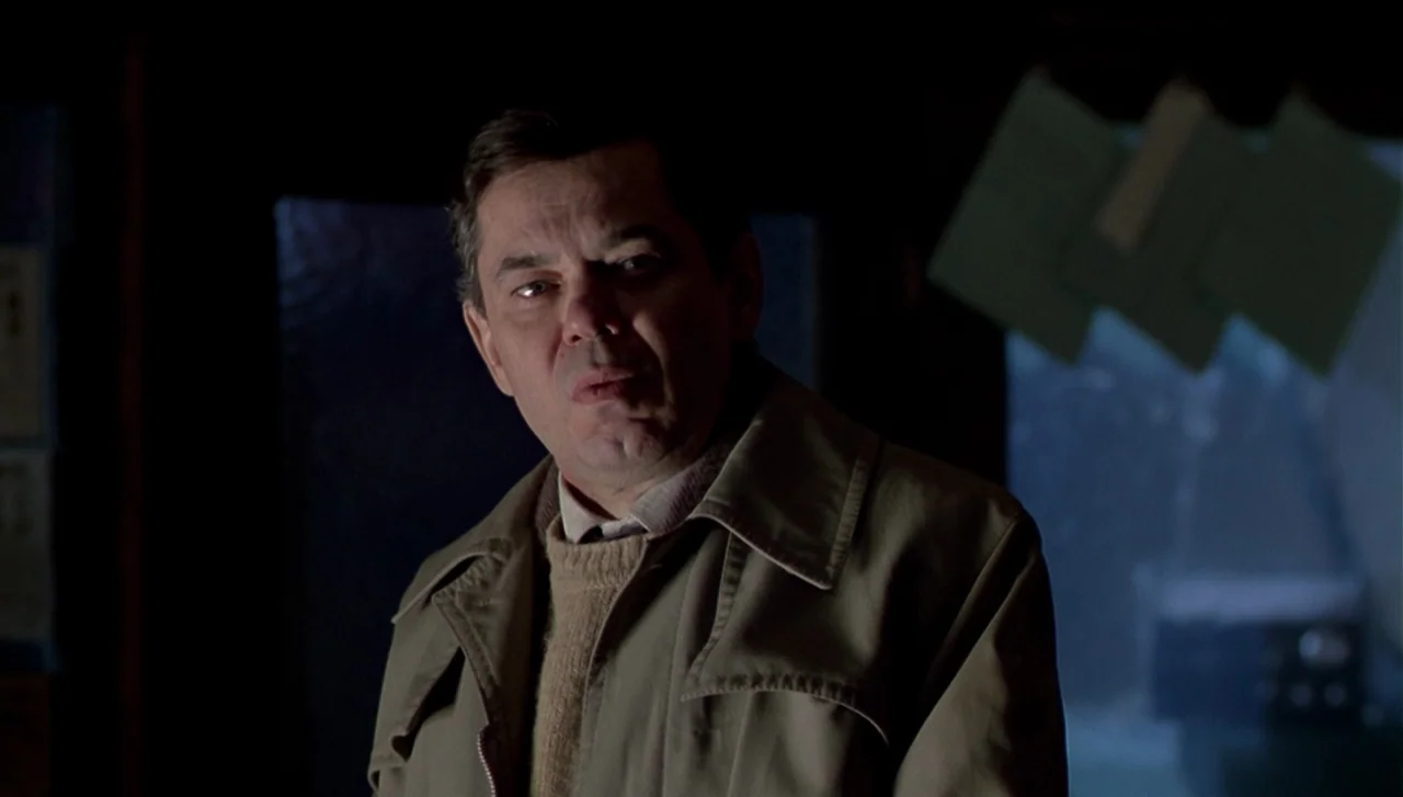 The Dead Zone (1983) movie still: medium — A dimly lit room with dark walls and a large window in the background. A middle-aged man in a tan jacket…