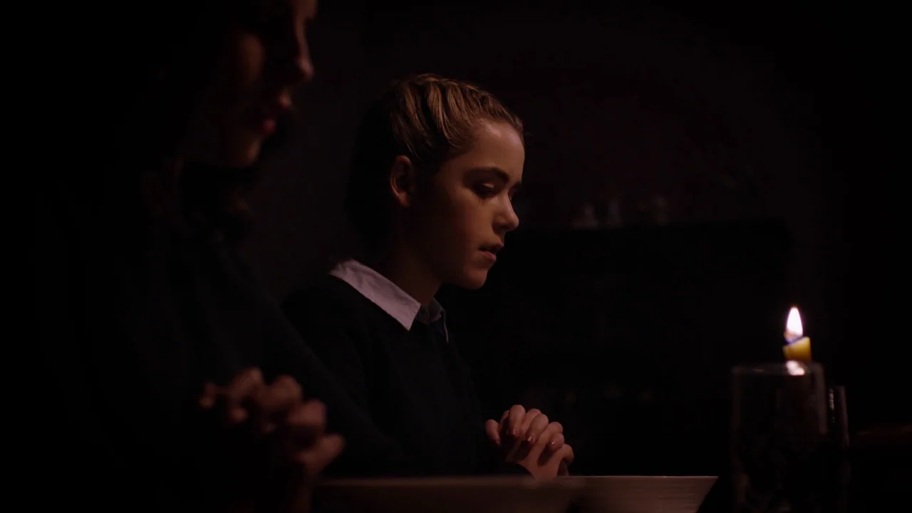 The Blackcoat’s Daughter (2015) movie still: medium — A darkened room features two people sitting at a table in near total darkness. The individuals…