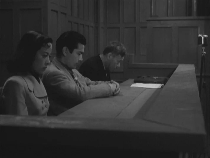 Scandal (1950) movie still: medium — A dark wooden room with paneled walls contains a long legal bench and papers. Three people sit in a row at the…
