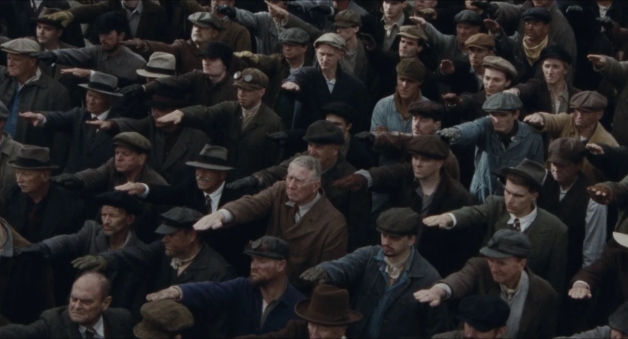 Origin (2023) movie still: wide — A dense gathering of men in period-appropriate work clothing stands in an open area. The crowd performs a unified…
