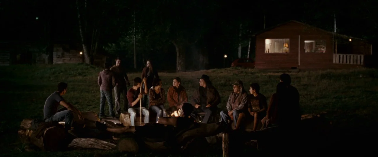 The Twilight Saga: Eclipse (2010) movie still: wide — A group of people gathers around a campfire in a dark, grassy clearing at night. A small lit…