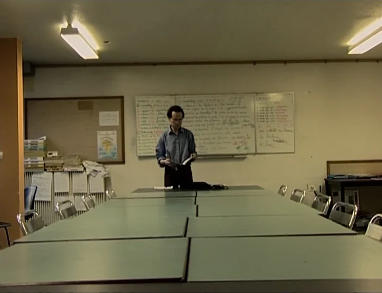 The Gleaners & I (2000) movie still: wide — A plain classroom features a long table in the foreground and whiteboards with handwritten text. A…