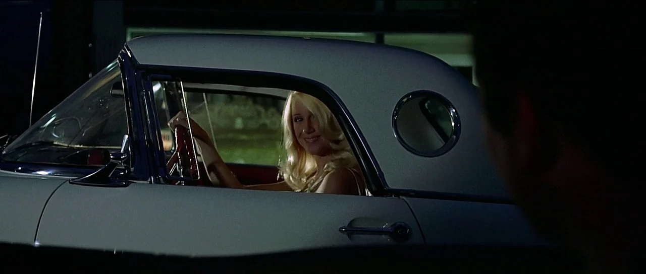 American Graffiti (1973) movie still: medium — A blonde woman sits in the driver seat of a vintage white convertible at night. She looks through the…