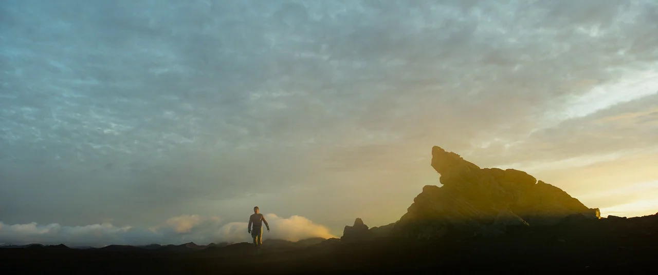 Oblivion (2013) movie still: wide — A vast, dark volcanic plain extends towards jagged rock formations under a cloudy sky. A solitary man walks…