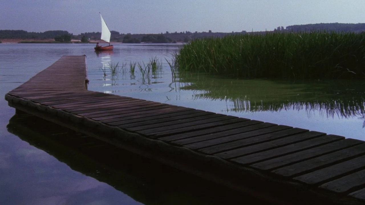 Insignificance (1985) movie still: wide — A wooden plank dock extends into a still, calm body of water. Reeds and marsh grass grow near the shore…
