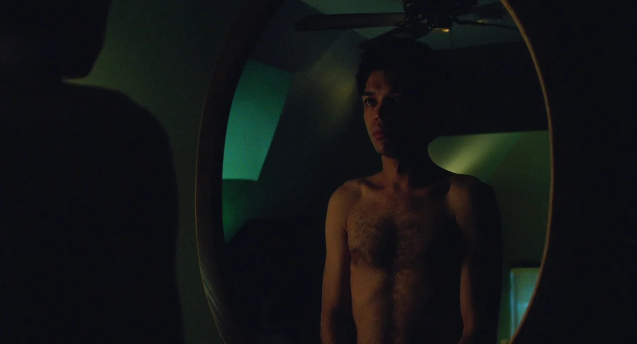 I Saw The TV Glow (2024) movie still: medium — A man stands before a circular wall mirror in a dimly lit dark-toned room. He stares forward at his…