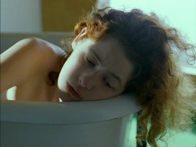 Helas Pour Moi (1993) movie still: closeup — A close view of a woman resting inside a white ceramic basin. She leans her face against the edge of…