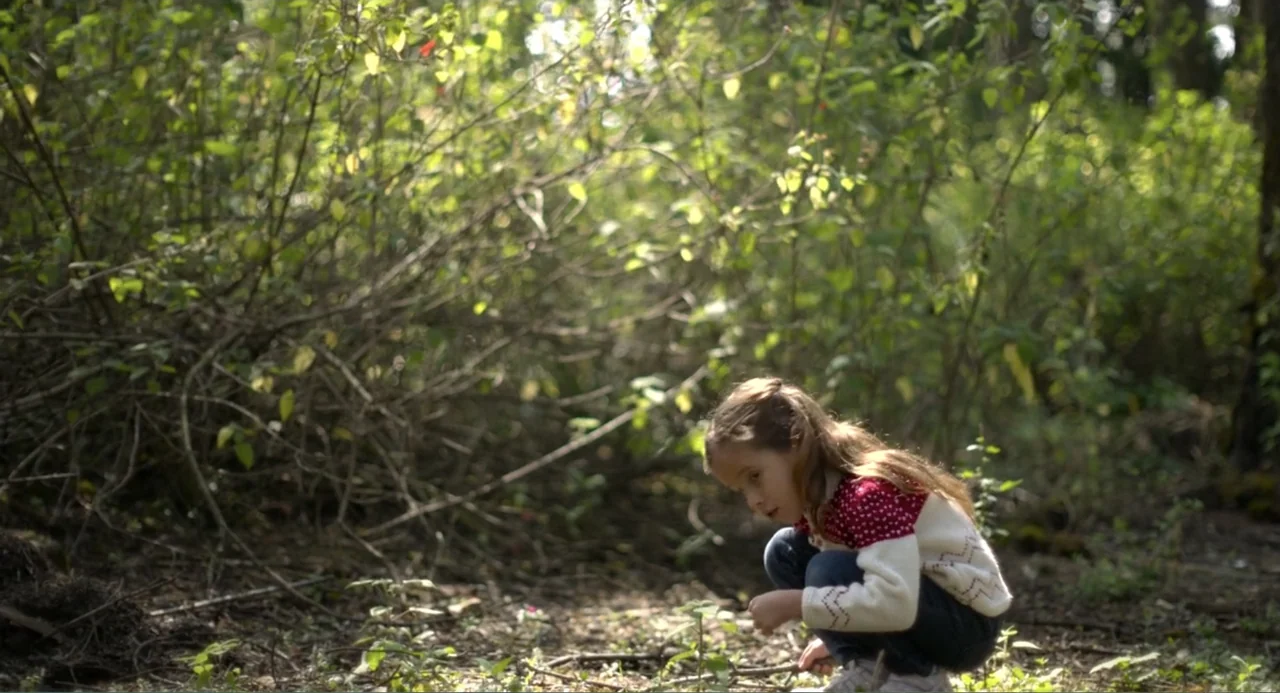 Heartbeat (2008) movie still: medium — A dense patch of wild woodland with thick undergrowth and dappled sunlight. A young girl with long brown hair…