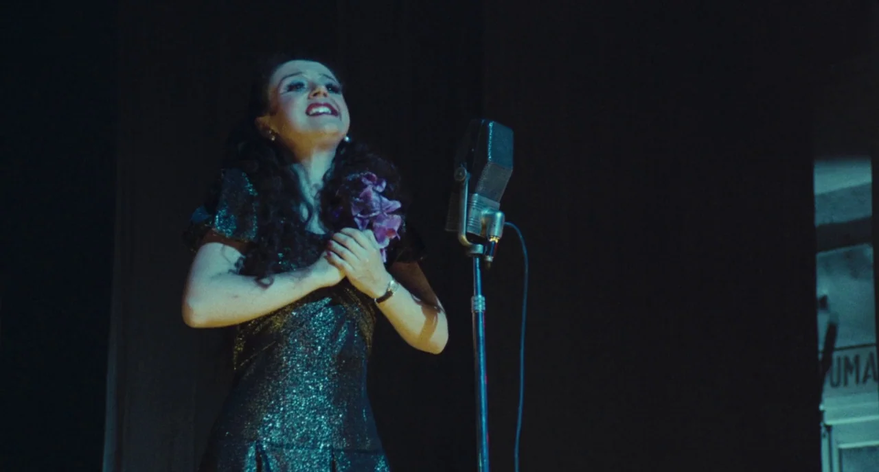 Roma (1972) movie still: medium — A woman stands alone on a dark stage before a tall vintage microphone. She clutches a purple flower while singing…