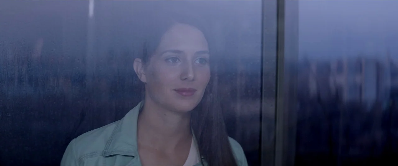 Loveless (2017) movie still: medium — A young woman stands behind a large pane of glass covered in fine raindrops. She looks outward toward an…
