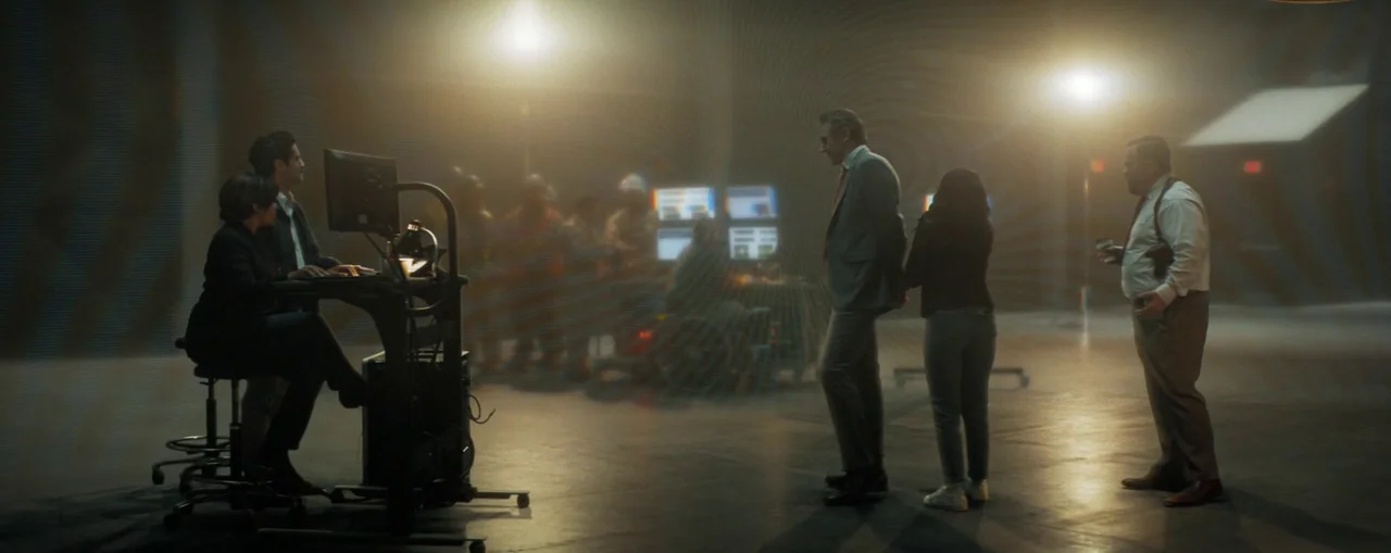 The Naked Gun (2025) movie still: wide — A dark, industrial command center with multiple monitors and blurred silhouettes in the background. A man…