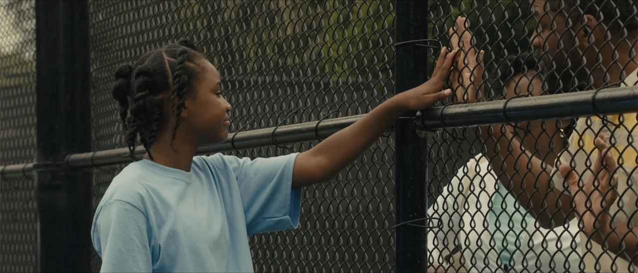 King Richard (2021) movie still: medium — A chain link fence separates two people standing outdoors during the day. A young girl on the left reaches…
