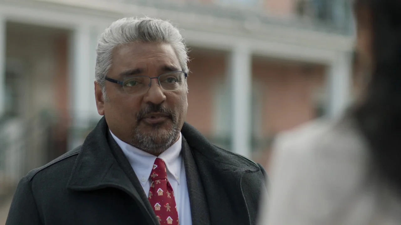 A Gingerbread Christmas (2022) movie still: medium — A man in a winter coat stands outside a building with white architectural columns. The man…