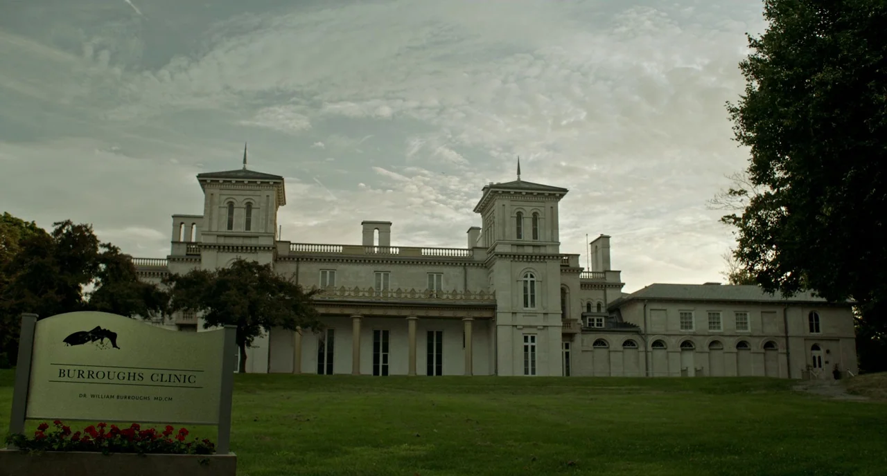 Rabid (2019) movie still: establishing — A large, pale stone mansion with symmetrical towers serves as the Burroughs Clinic building. A sign in the…