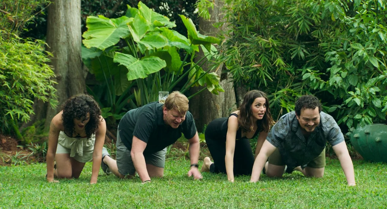 Deep Water (2022) movie still: fullbody — Four people crawl on a green grass lawn backed by dense tropical foliage. A glass of liquid sits on the…