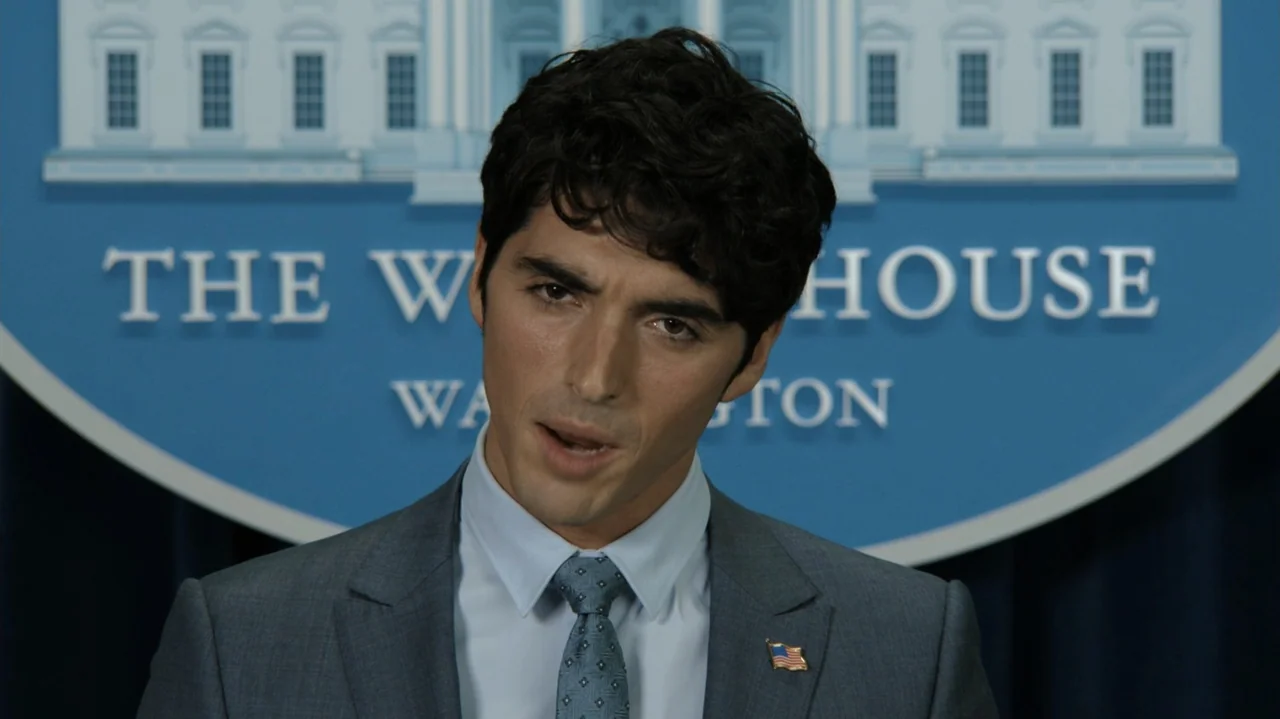 Red, White & Royal Blue (2023) movie still: medium — A man stands in front of a blue White House press briefing backdrop. He speaks while wearing a…