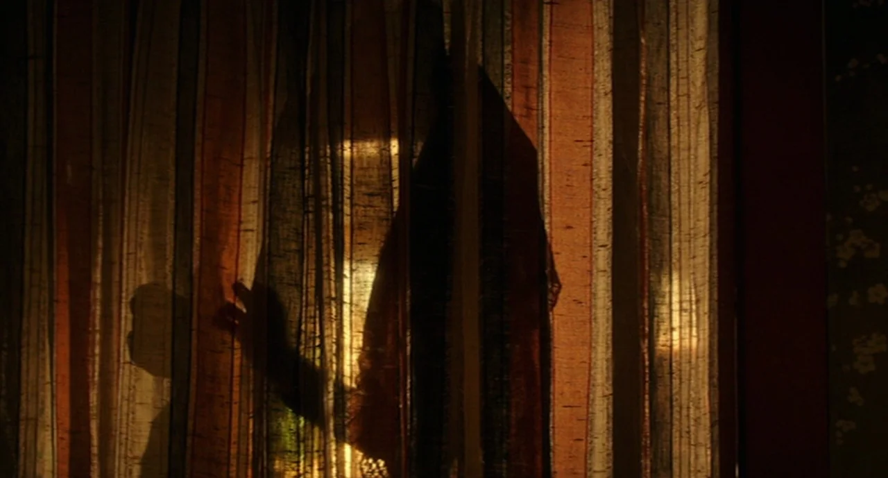 Ouija: Origin of Evil (2016) movie still: closeup — A close-up of amber and brown striped curtains backlit by bright daylight. A dark silhouette of…